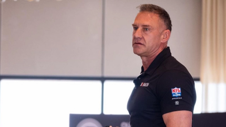 Man in black NFL Alumni polo shirt giving a presentation in a bright, modern conference room.