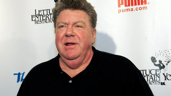 George Wendt posing at a red-carpet event in a black polo shirt and smiling for the camera.