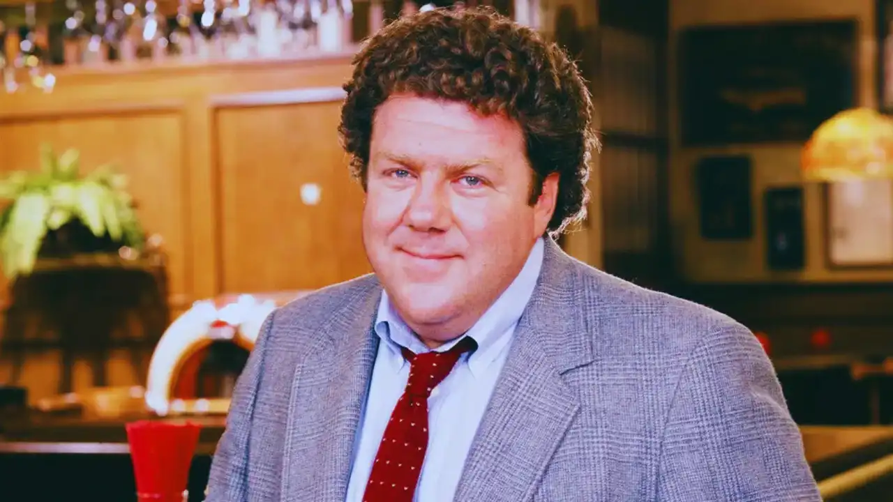 George Wendt as Norm Peterson smiling at the Cheers bar in a gray suit and red tie.