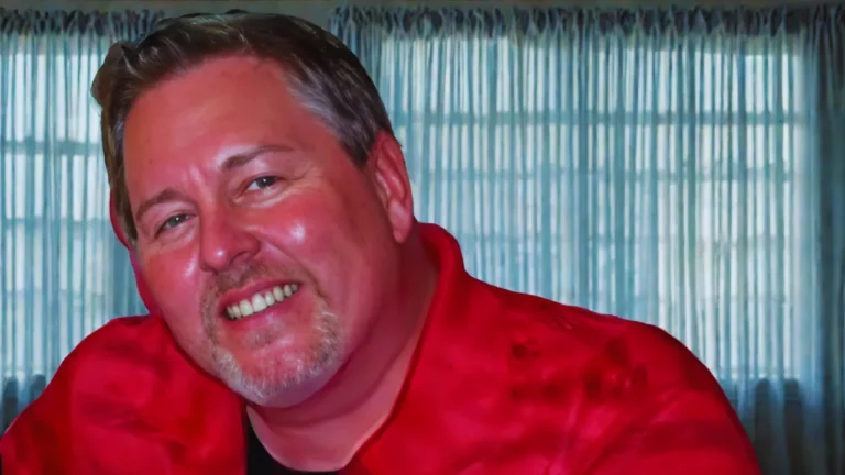 Smiling portrait of Jake Merendino wearing a red shirt, in front of window curtains.