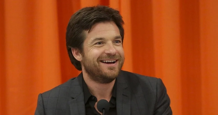 Jason Bateman smiling during a public appearance, wearing a dark suit, featured in net worth article.