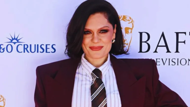 Jessie J at BAFTA event in burgundy suit and striped tie, smiling on red carpet.