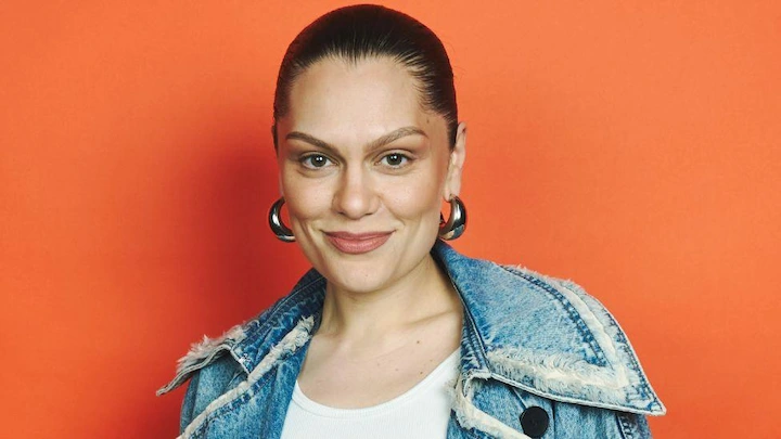 Jessie J smiles in denim jacket with orange background after announcing breast cancer diagnosis.