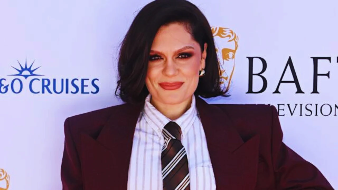 Jessie J at BAFTA event in burgundy suit and striped tie, smiling on red carpet.