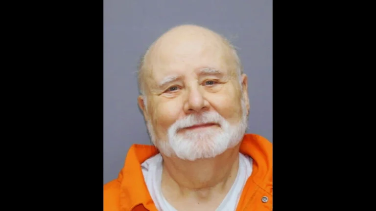 Mugshot of John Edward Robinson in an orange jumpsuit, with a white beard and slight smile.