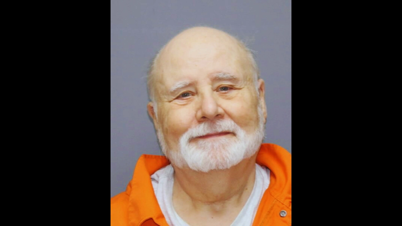 Mugshot of John Edward Robinson in an orange jumpsuit, with a white beard and slight smile.