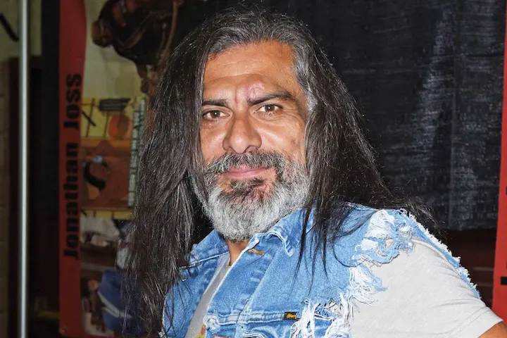 Jonathan Joss at a public event, wearing a denim vest, smiling, with long hair and a graying beard.