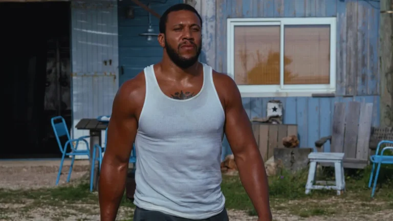 Bastien stands sweaty in a white tank top outside a rustic building, muscles tense and expression resolute.