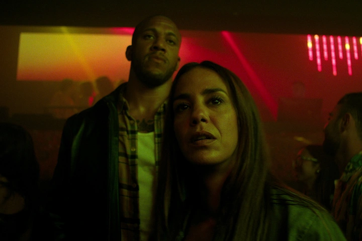 Bastien and Emma in a crowded neon nightclub, their faces lit by red and green lights, tension in their stance.