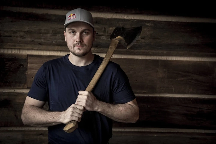 Matt Cogar holding an axe while wearing a Red Bull cap in a rustic wooden setting
