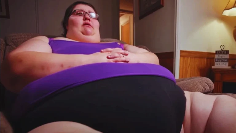 Megan Davis seated on a couch wearing purple top and glasses with hands resting on her belly warmly.