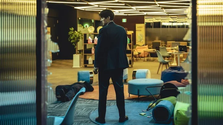 Nam Gi-jun stands in a sleek hotel lounge amid overturned furniture after a violent event.