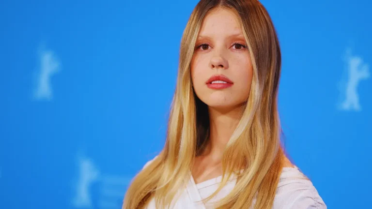 Mia Goth at a film festival press event in front of a blue backdrop, wearing a white top and makeup.