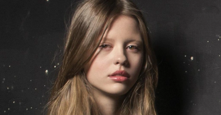 Close-up headshot of Mia Goth with parted blonde hair, natural freckles, and a thoughtful, direct stare.