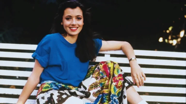 Mia Sara smiling on a bench in a blue top and colorful skirt from Ferris Bueller’s Day Off era.