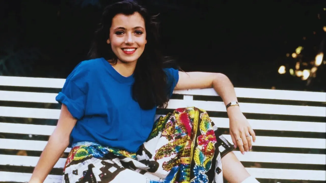 Mia Sara smiling on a bench in a blue top and colorful skirt from Ferris Bueller’s Day Off era.