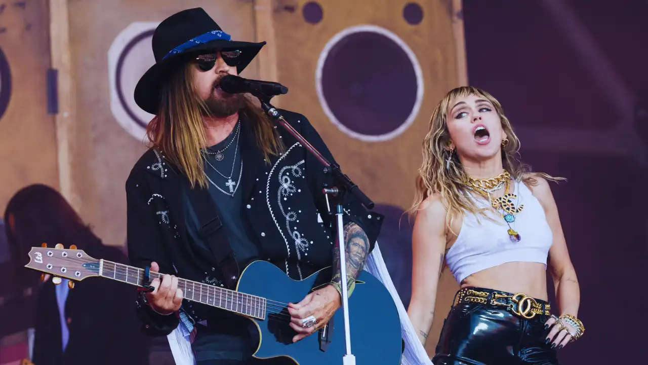Billy Ray Cyrus and Miley Cyrus perform together on stage with guitar and dynamic stage presence.