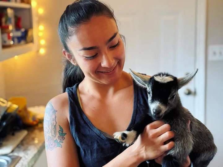 Alexia Huffman holding a baby goat inside a rustic kitchen, smiling as she cares for livestock.