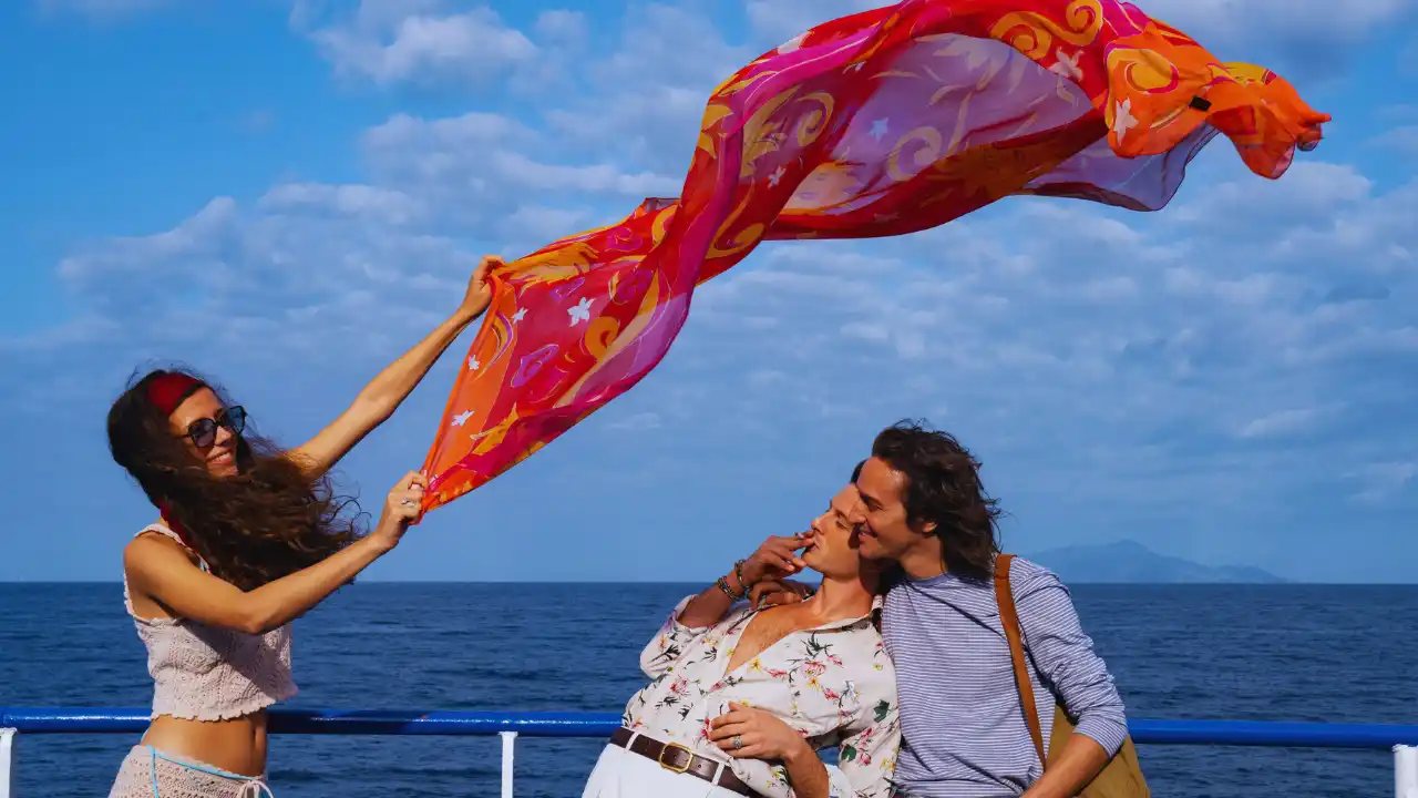 Colorful scarf blowing in wind as three young adults laugh on a boat under a bright blue sky.