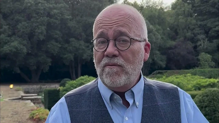 Man in a blue shirt and vest standing outdoors with lush green trees and garden in the background.