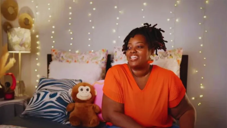 Rhasha Newkirk smiling in cozy bedroom with fairy lights and stuffed monkey on bed.