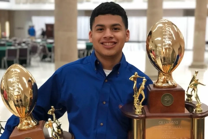Rudy Peña smiles proudly holding football trophies at school event.