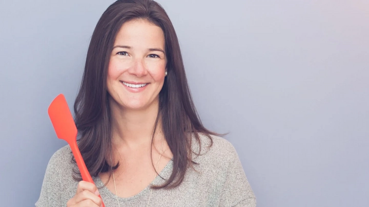 Samantha Rose smiling and holding a red spatula, wearing a gray sweater, against a gray background.