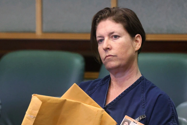 Sarah Boone sits in a courtroom wearing a dark jumpsuit, holding documents, with stern expression...
