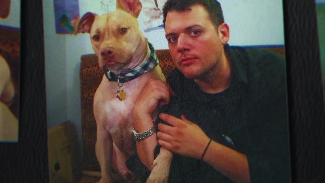 Anthony Strangis aka Shane Fox kneels beside a tan pitbull dog wearing a plaid collar at home.