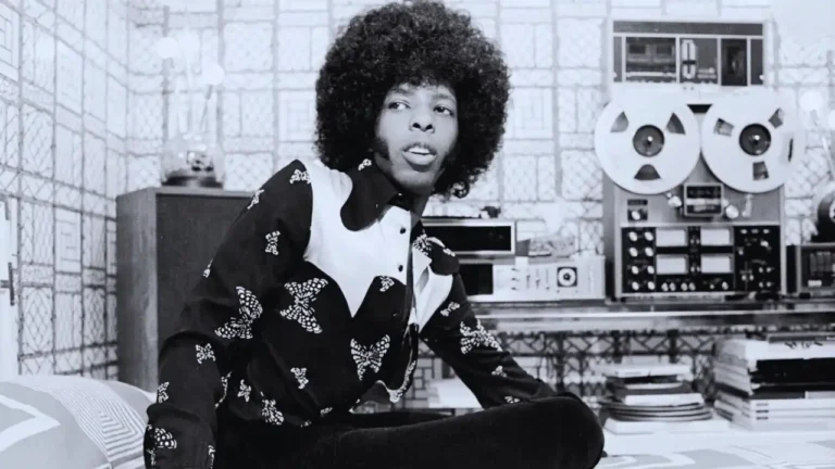 Sly Stone in a recording studio wearing patterned shirt with large tape reels behind him.