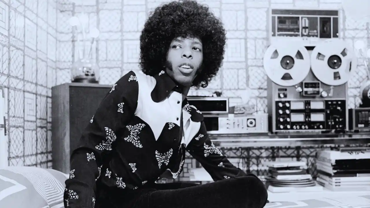 Sly Stone in a recording studio wearing patterned shirt with large tape reels behind him.