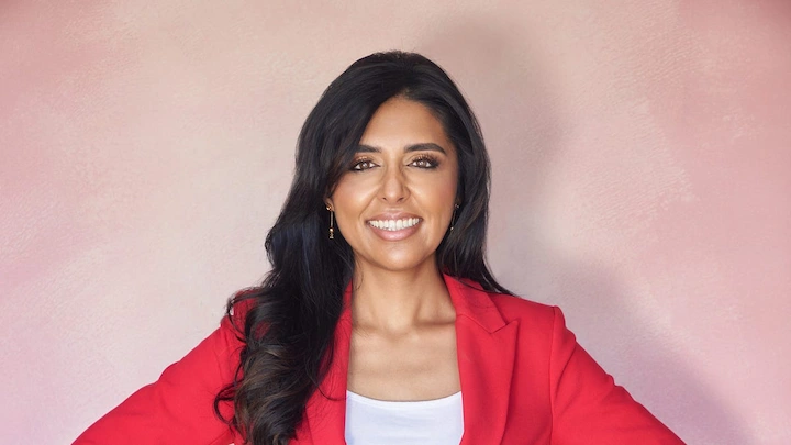 Confident woman in red blazer smiling against soft pink wall with hands on hips.