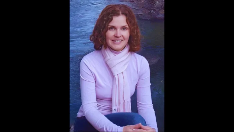 Tara Lynn Grant smiling in a light pink top and scarf, seated by a riverbank with calm water behind.