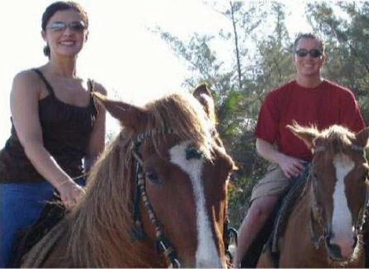 Tara Lynn Grant and her husband Stephen riding horses on a sunny tropical beach, both smiling.