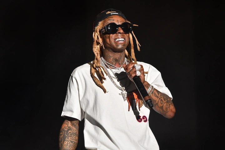 Lil Wayne smiling on stage in sunglasses and white t-shirt during his 2025 Tha Carter VI Tour.