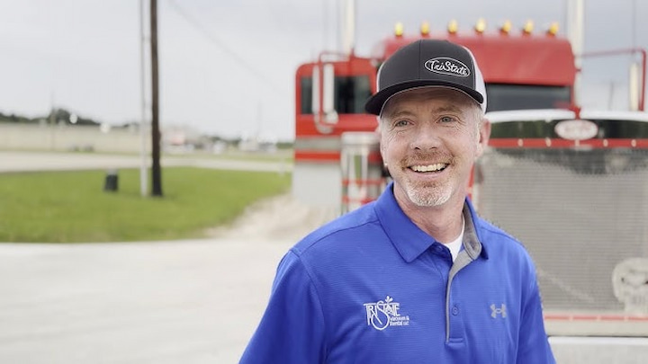 Troy Massey smiling in front of a red truck, wearing a blue shirt and black cap, showcasing success