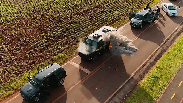 Aerial shot of armored van smoking on a rural road flanked by three black SUVs during a staged heist.