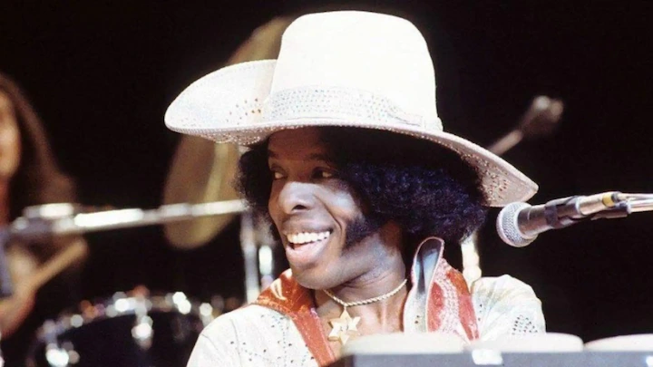 Sly Stone smiling on stage wearing a wide-brim hat during a live keyboard performance.