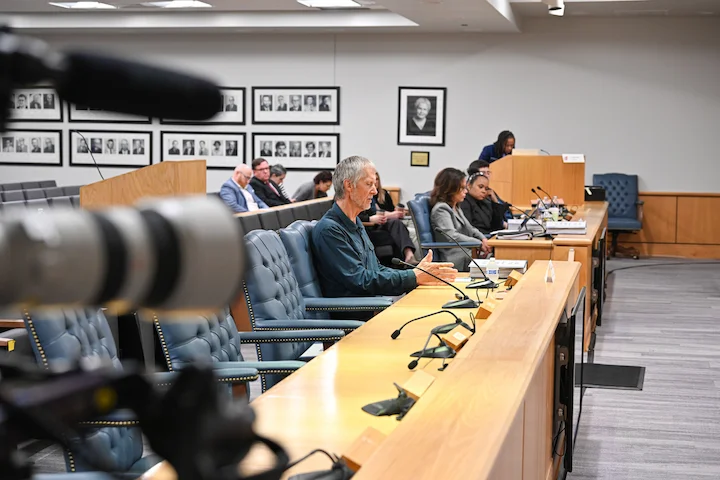 Tym Catterson seated at hearing table, camera lens in foreground, hearing room backdrop behind him.!