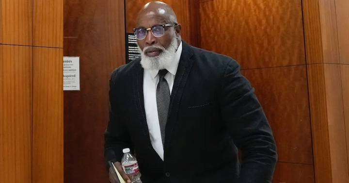 Renard Spivey in court wearing a suit and glasses, holding a water bottle and book during trial
