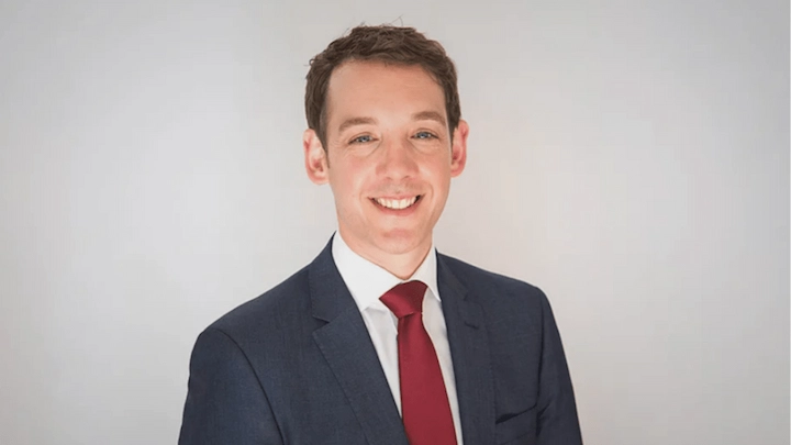 Steve Dimopoulos wearing a navy suit and red tie, smiling in a professional studio portrait